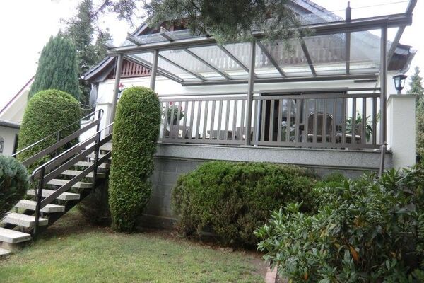 Überdachte Terrasse mit Glasdach und Treppe, umgeben von gepflegten Gartensträuchern.