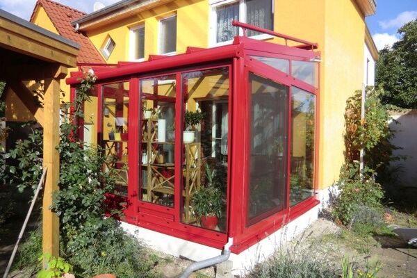 Moderner Wintergarten mit roten Aluminiumrahmen an gelbem Einfamilienhaus im Garten.
