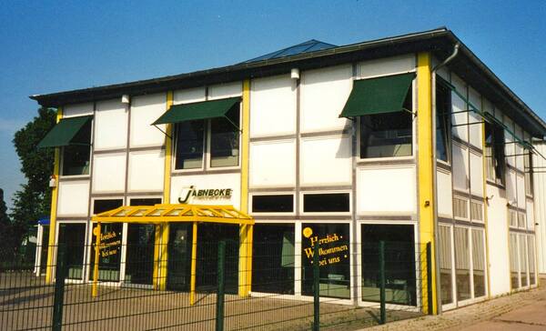 Modernes, zweistöckiges Firmengebäude mit gelbem Eingangsbereich und begrünten Außenjalousien.